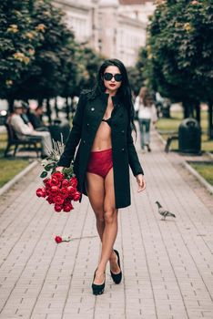 Elegant beautiful brunette woman in red shorts and black coat posing on a city streets with a big bouquet of red roses. Birthday. Valentines day.