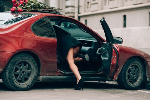 Elegant beautiful brunette woman in red shorts and black coat posing near red car. big bouquet of red roses on a car roof. Birthday. March 8.