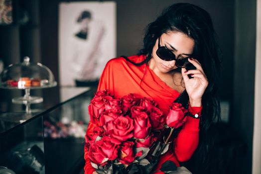 Charming young woman in red sexy dress and sunglasses posing with a bouquet of red roses. photo of a seductive woman with black hair. Selective focus, filmgrain.