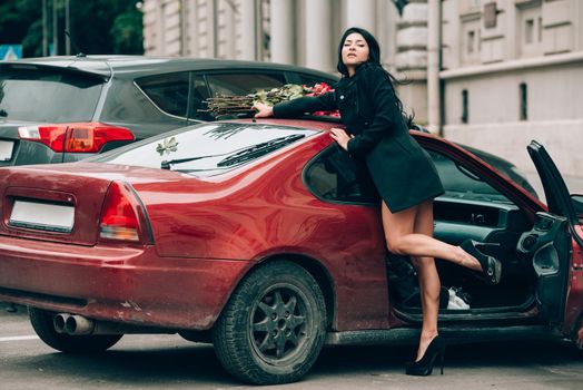 Elegant beautiful brunette woman in red shorts and black coat posing near red car. big bouquet of red roses on a car roof. Birthday. March 8.