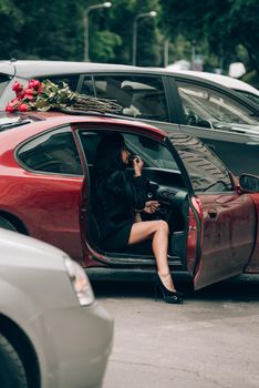 Elegant beautiful brunette woman in red shorts and black coat posing near red car. big bouquet of red roses on a car roof. Birthday. March 8.