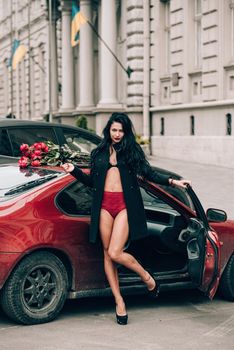 Elegant beautiful brunette woman in red shorts and black coat posing near red car. big bouquet of red roses on a car roof. Birthday. March 8.