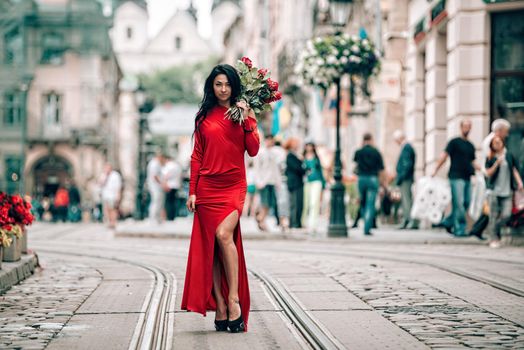 Charming young woman in red sexy dress posing with a bouquet of red roses. photo of a seductive woman with black hair on the city streets. Selective focus, filmgrain.