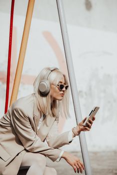 Portrait of blonde hipster fashion model wear stylish wireless headphones enjoy listen new cool music. woman wearing white jacket, blouse, tights and brown leather skirt