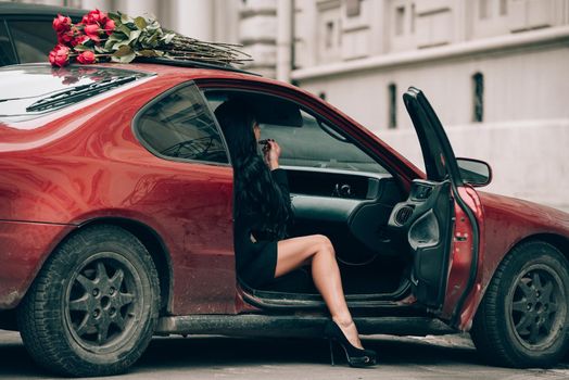 Elegant beautiful brunette woman in red shorts and black coat posing in a red car. big bouquet of red roses on a car roof. Birthday. March 8. Valentines day.