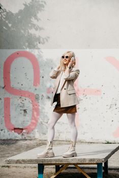 Portrait of blonde woman in headphones and sunglasses listening music. woman wearing white jacket, blouse, tights and brown leather skirt. white high leather boots