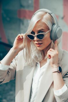 Portrait of blonde hipster fashion model wear stylish wireless headphones enjoy listen new cool music. woman wearing white jacket, blouse, tights and brown leather skirt