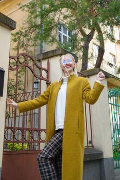 Fashionable beautiful young woman with blond hair in a stylish long coat, checkered pants and glasses poses in the city streets. Feminine urban style.