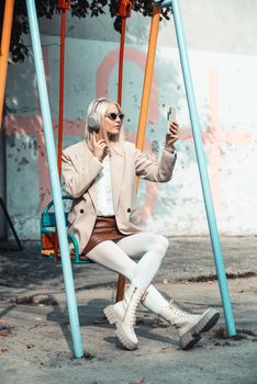 Young smiling cheerful woman outdoors using social media apps on phone for video chatting and stying connected. woman wearing white jacket, blouse, tights and brown leather skirt