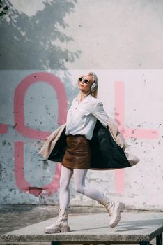 Portrait of blonde woman in headphones and sunglasses listening music. woman wearing white jacket, blouse, tights and brown leather skirt. white high leather boots