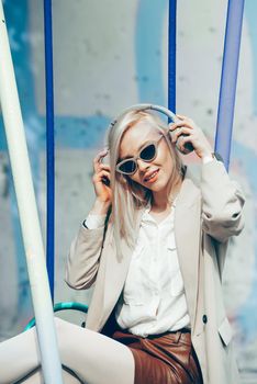 Portrait of blonde hipster fashion model wear stylish wireless headphones enjoy listen new cool music. woman wearing white jacket, blouse, tights and brown leather skirt