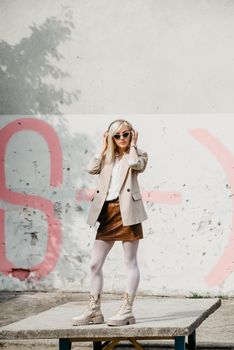 Portrait of blonde woman in headphones and sunglasses listening music. woman wearing white jacket, blouse, tights and brown leather skirt. white high leather boots