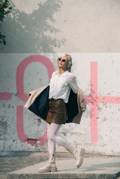 Portrait of blonde woman in headphones and sunglasses listening music. woman wearing white jacket, blouse, tights and brown leather skirt. white high leather boots