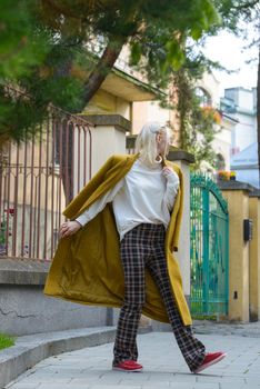 Fashionable beautiful young woman with blond hair in a stylish long coat, checkered pants, red shoes and glasses poses in the city streets. Feminine urban style.