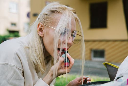 young smiling hipster happy woman in glasses doing make-up using lipstick and mirror outdoors