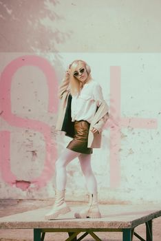 Portrait of blonde woman in headphones and sunglasses listening music. woman wearing white jacket, blouse, tights and brown leather skirt. white high leather boots