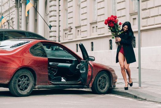 Elegant beautiful brunette woman in red shorts and black coat posing near red car. big bouquet of red roses. Birthday. March 8.