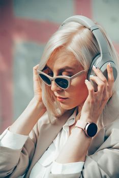 Portrait of blonde hipster fashion model wear stylish wireless headphones enjoy listen new cool music. woman wearing white jacket, blouse, tights and brown leather skirt