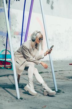 Portrait of blonde hipster fashion model wear stylish wireless headphones enjoy listen new cool music. woman wearing white jacket, blouse, tights and brown leather skirt