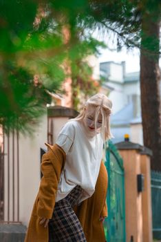 Fashionable beautiful young woman with blond hair in a stylish long coat, checkered pants, red shoes and glasses poses in the city streets. Feminine urban style.