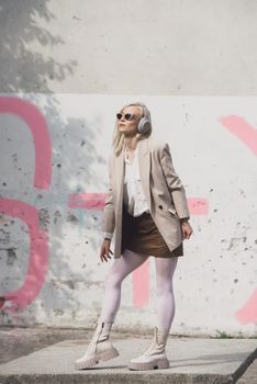 Portrait of blonde woman in headphones and sunglasses listening music. woman wearing white jacket, blouse, tights and brown leather skirt. white high leather boots