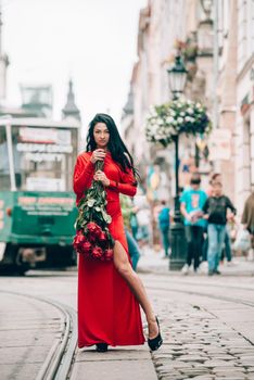 Charming young woman in red sexy dress posing with a bouquet of red roses. photo of a seductive woman with black hair on the city streets. Selective focus, filmgrain.