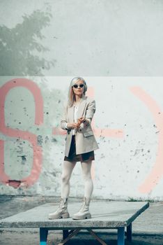 Portrait of blonde woman in headphones and sunglasses listening music. woman wearing white jacket, blouse, tights and brown leather skirt. white high leather boots