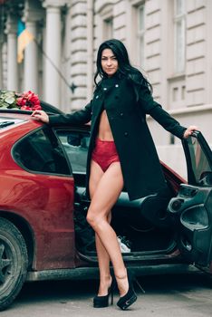 Elegant beautiful brunette woman in red shorts and black coat posing near red car. big bouquet of red roses on a car roof. Birthday. March 8.