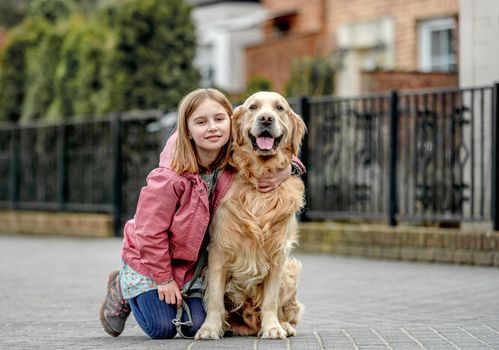 Preteen girl hugging golden retriever dog outdoors. Pretty child kid with purebred dog labrador sit on the ground happy
