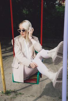 Portrait of blonde hipster fashion model wear stylish wireless headphones enjoy listen new cool music. woman wearing white jacket, blouse, tights and brown leather skirt