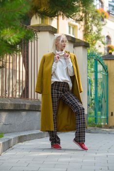 Fashionable beautiful young woman with blond hair in a stylish long coat, checkered pants, red shoes and glasses poses in the city streets. Feminine urban style.