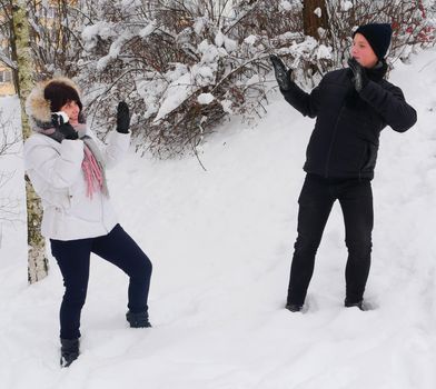 Mom in a white jacket and scarf plays snowballs with her teenage son in a black jacket, hat, trousers against the backdrop of snow.