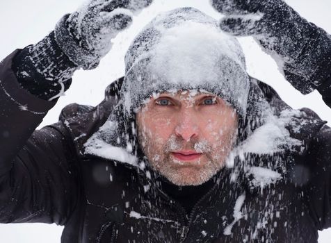 face of a handsome man with a beard in bad weather conditions in winter. Portrait of a climber during a storm. A man in black gloves raises his hands in the snow.