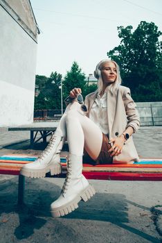 Portrait of blonde hipster fashion model wear stylish wireless headphones enjoy listen new cool music. woman wearing white jacket, blouse, tights and brown leather skirt