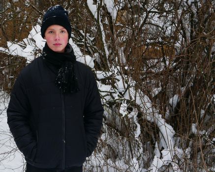 A waist-length portrait of a cute teen boy in a black hat, scarf, jacket holds his hands in his pockets against the backdrop of a snowy bush.