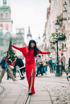 Charming young woman in red sexy dress posing with a bouquet of red roses. photo of a seductive woman with black hair on the city streets. Selective focus, filmgrain.