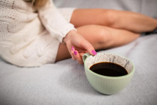 Woman holds coffee cup close to her. Slender female legs with pale complexion. Relaxing in the comfort of home.