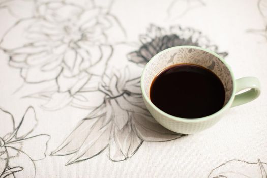 View from above. On the table in the room at home stands a porcelain cup with hot aromatic coffee. A tablecloth with a floral pattern.