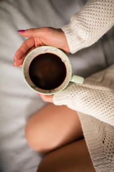 Close-up frame. A woman with slim legs sits on a bed in a living room and next to her holds a porcelain cup with hot coffee.