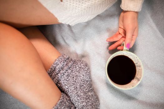 Slender female legs in grey wool socks. Porcelain cup with coffee in woman's hands. Relaxing in the comfort of home.