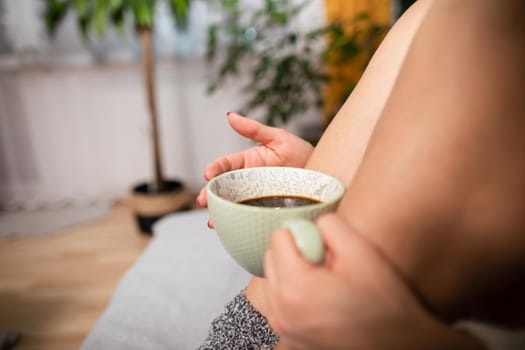 Woman holds coffee cup close to her. Slender female legs with pale complexion. Relaxing in the comfort of home.