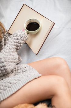 A woman lies on a bed with a book and a cup of coffee. Neat female legs with light complexion of skin. Aromatic coffee time while relaxing.
