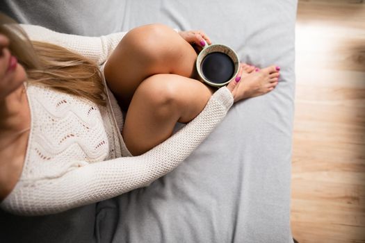 Cup of morning coffee held by female hand. Sexy female legs with light skin complexion. Relaxation in the comfort of your home.