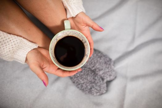 Porcelain cup with coffee held by woman's hands. Neat female legs. Relaxation in the comfort of home.