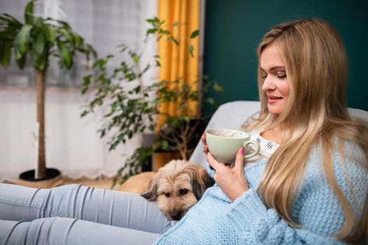 A young woman is sitting on the sofa with her dog and holding a cup of coffee. A moment of relaxation spent with her pet.