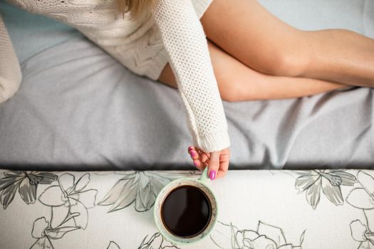 Young woman with long shapely legs is sitting on sofa at home. Porcelain cup with aromatic coffee held by woman's hand.