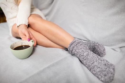 Woman's shapely legs dressed in warm woolen socks. Porcelain cup with coffee held by feminine hands. Relaxation in the comfort of your home.