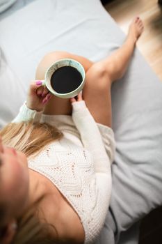 Cup of morning coffee held by female hand. Sexy female legs with light skin complexion. Relaxation in the comfort of your home.