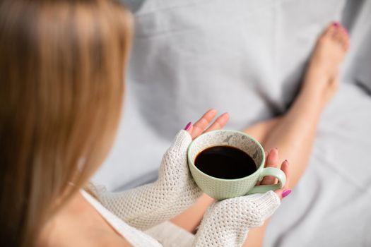A woman with blonde hair sits on a sofa and holds a cup of coffee in her hands. Neat legs with light complexion of skin. Love for coffee and relaxation.