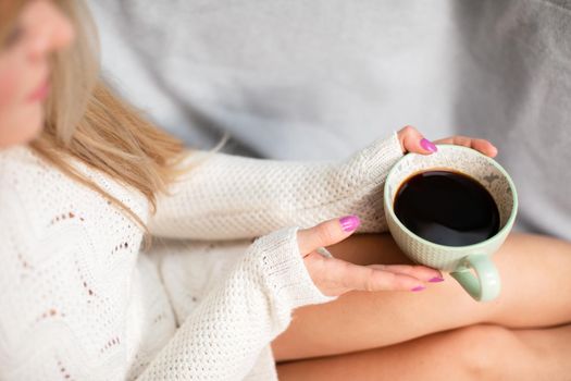 A woman with blonde hair sits on a sofa and holds a cup of coffee in her hands. Neat legs with light complexion of skin. Love for coffee and relaxation.